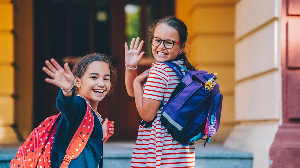 Two girls wearing backpacks waving