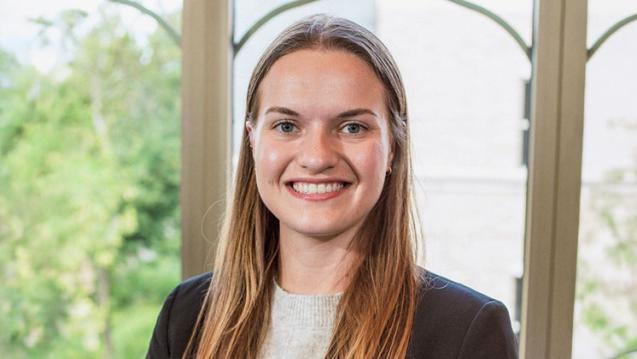 Elaina smiling wearing a navy blazer and gray shirt in front of window