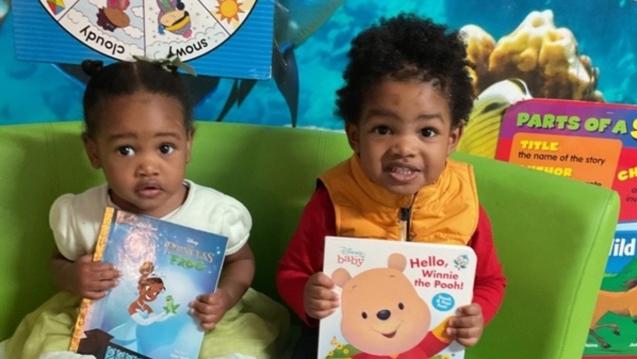 A toddler girl and a toddler boy sit on a sofa holding books