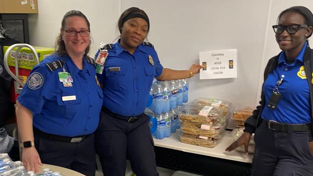 Three TSA agents standing by tables filled with water and food