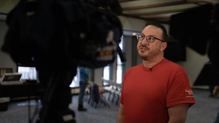 Behind the scenes photo of man speaking into camera in a red shirt