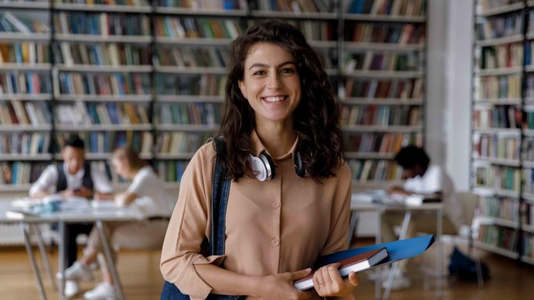 Young woman standing in a library with a shoulder bag and book in her hand