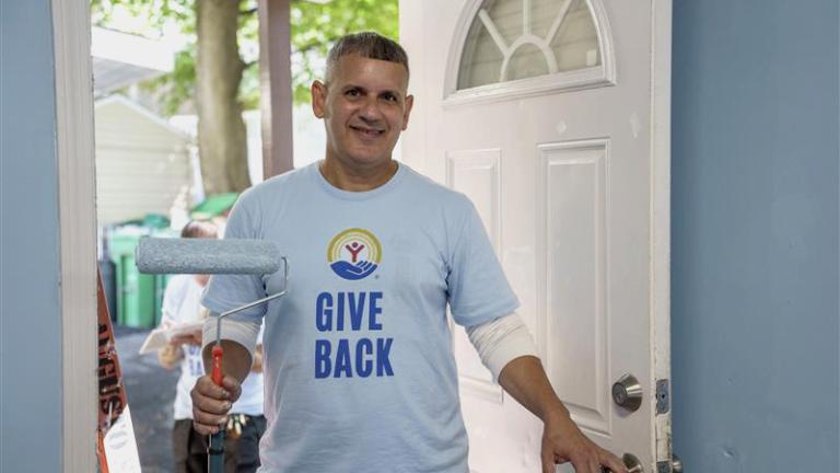 Man in a United Way shirt standing in the doorway of a home, holding a paint roller.