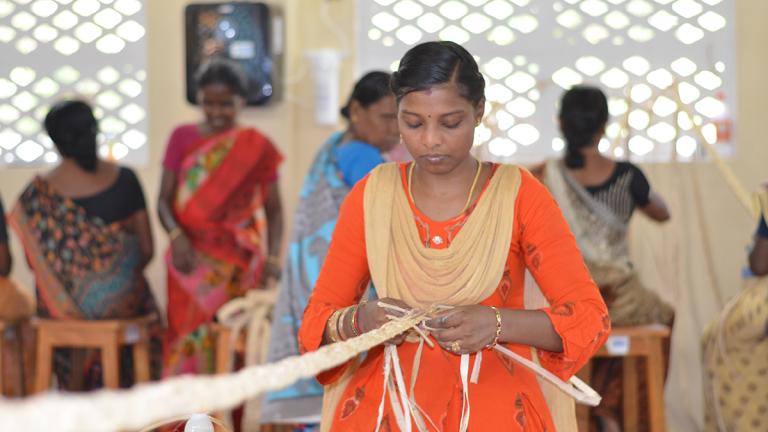 A woman wearing an orange sari weaves banana fiber.