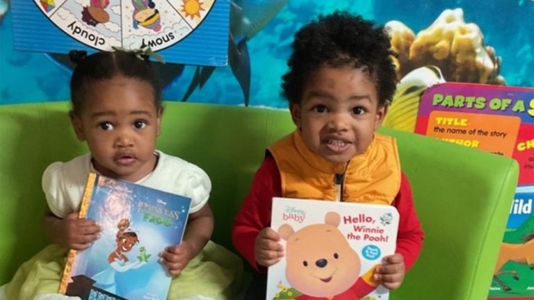 A toddler girl and a toddler boy sit on a sofa holding books
