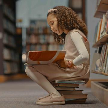 Child sitting on a stack of books reading a book in a library.