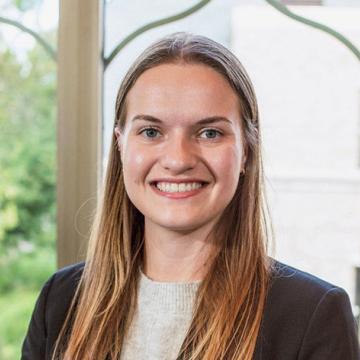 Elaina smiling wearing a navy blazer and gray shirt in front of window