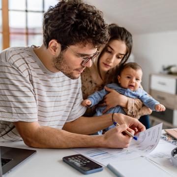 A man huddling over a bill while a woman and baby look over his shoulder 