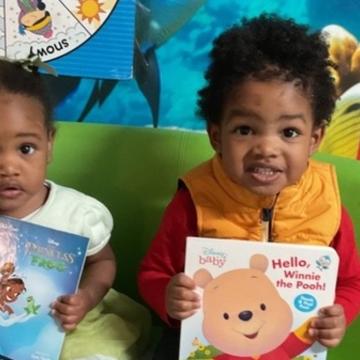 A toddler girl and a toddler boy sit on a sofa holding books