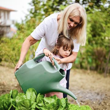 A woman standing with a young girl watering a plant together in a garden