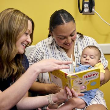 A mom with a baby on her laps sits with a medical professional who is turning the page of a book. 