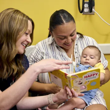 A mom with a baby on her laps sits with a medical professional who is turning the page of a book. 